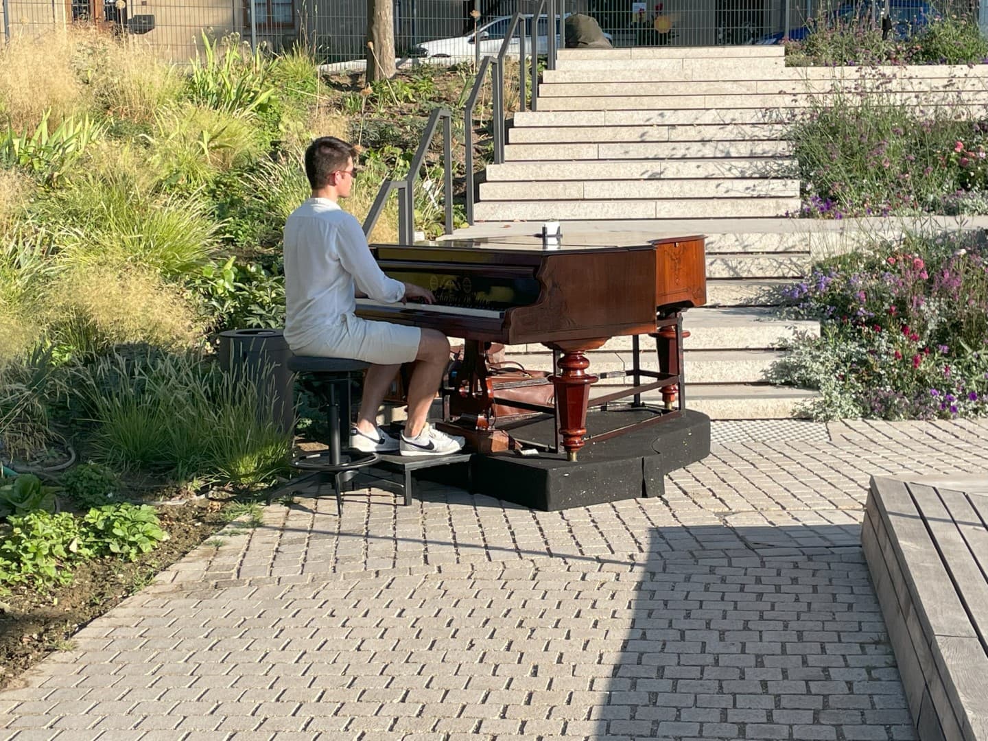 Ein junger Mann spielt an einem Klavier auf einem gepflasterten Platz zwischen Staudenbeeten