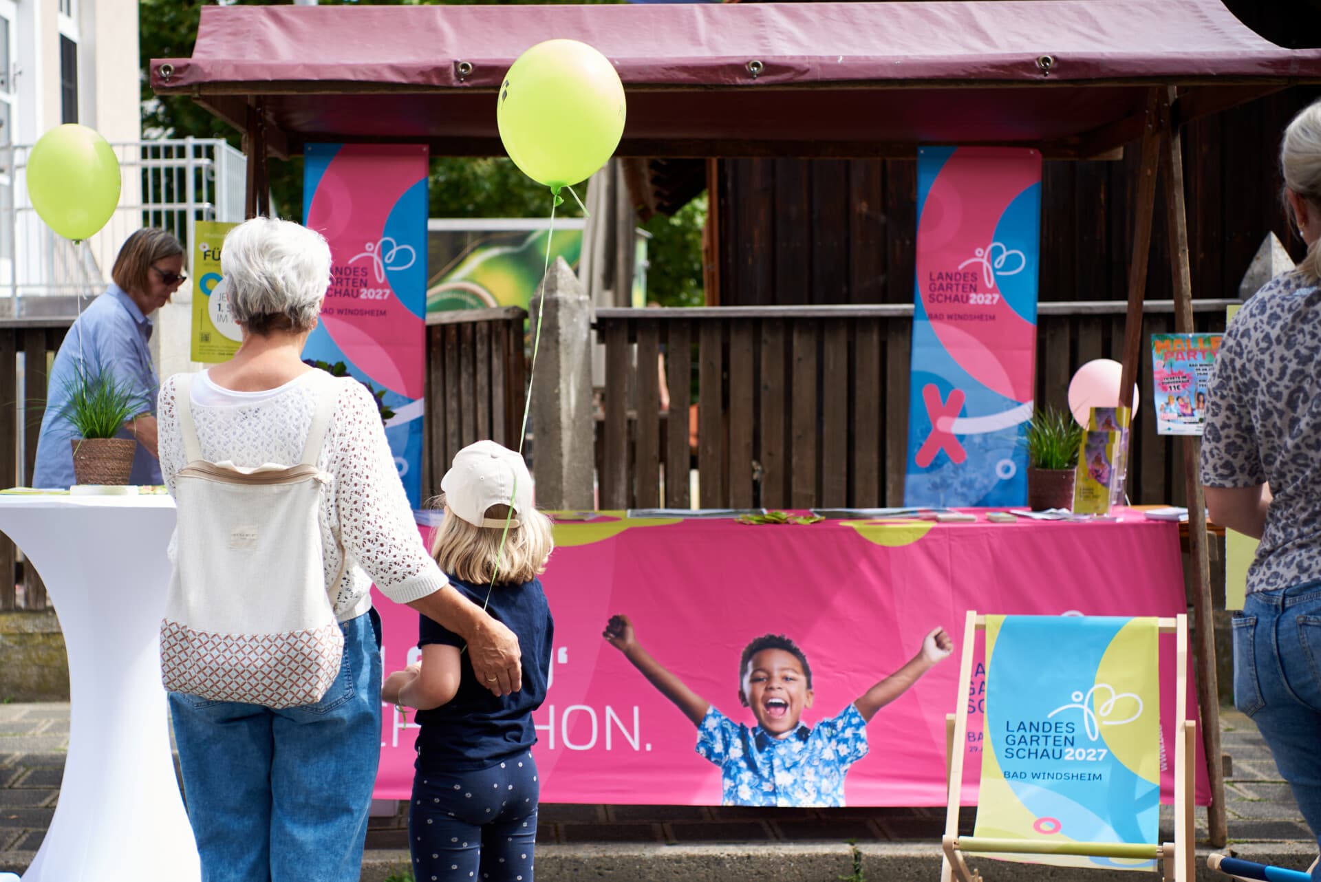 Infostand der Landesgartenschau auf dem Heimatfest, Besucherinnen mit Luftballons vor einem pinken Banner