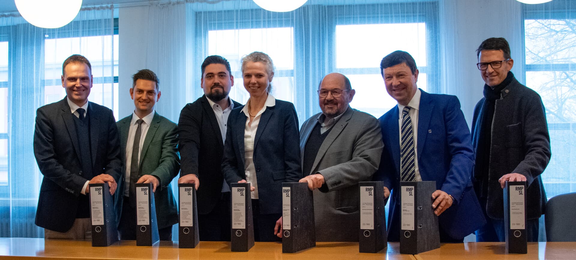 Sieben Personen stehen mit Aktenordnern an einem Konferenztisch