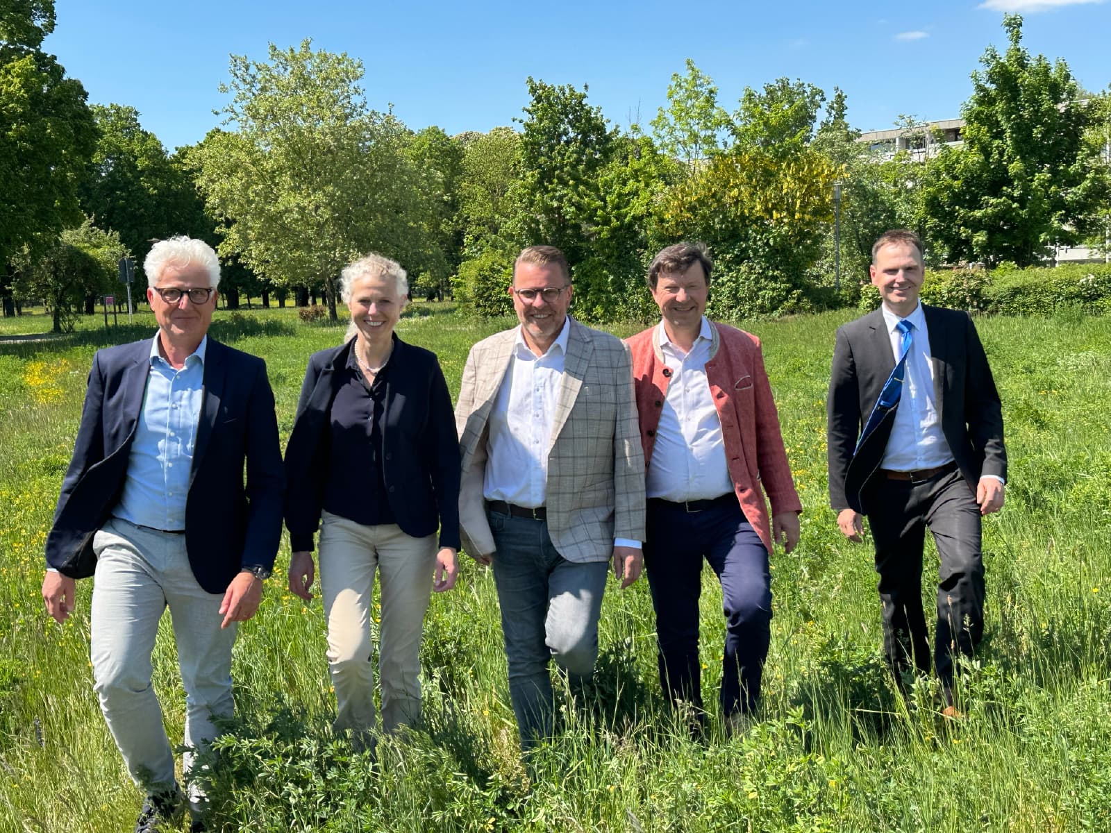 Fünf Personen in Anzügen gehen nebeneinander über eine Wiese