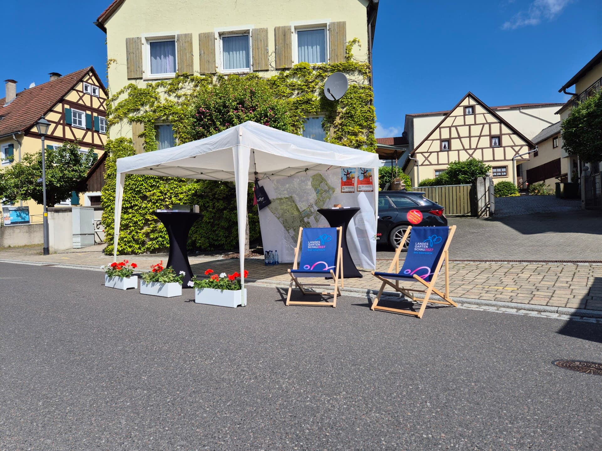 Weißer Pavillonstand der Landesgartenschau mit Liegestühlen in einer Dorfstraße