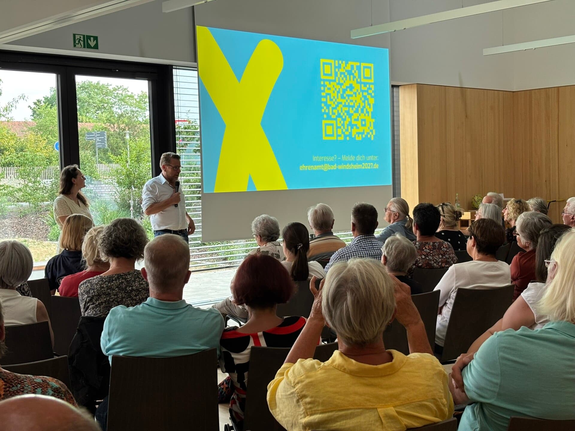 Voll besetzter Saal beim Infoabend, vorne zwei Personen vor einer Leinwand mit gelbem X und QR-Code